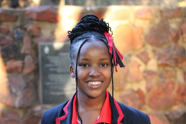 A learner in school blazer smiling in front of a stone wall
